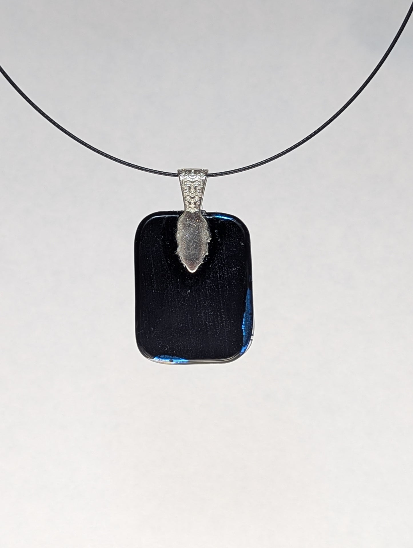 The black back of a blue dichroic fused glass pendant with a silver metallic clasp, displayed on a black stainless steel necklace.