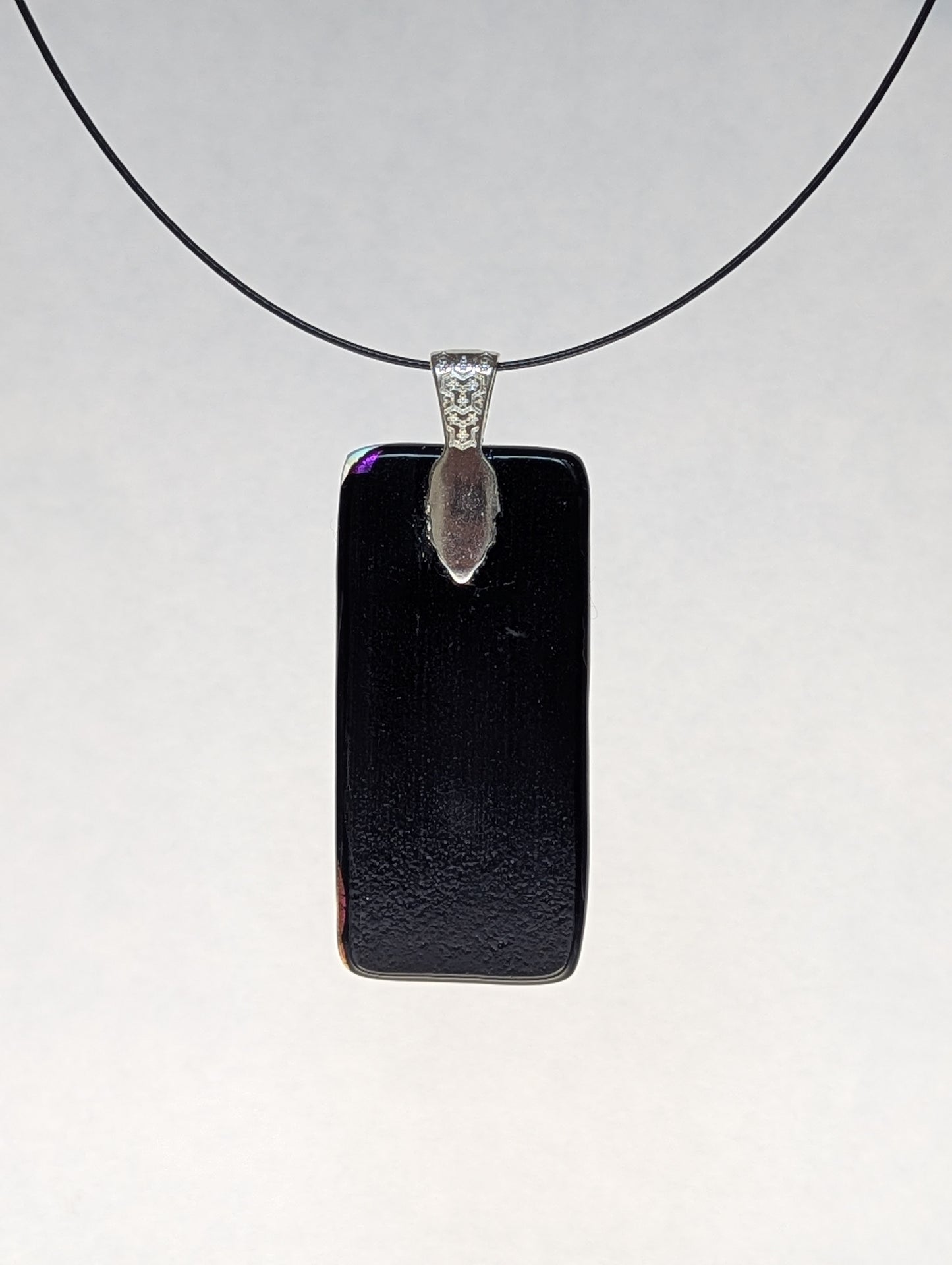 The black back of a purple dichroic fused glass pendant with a silver metallic clasp, displayed on a black stainless steel necklace.