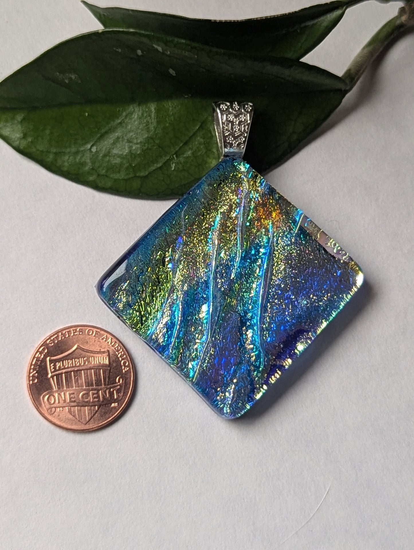A blue iridescent fused glass pendant with a silver metallic clasp, displayed on a leaf next to a penny for size reference.