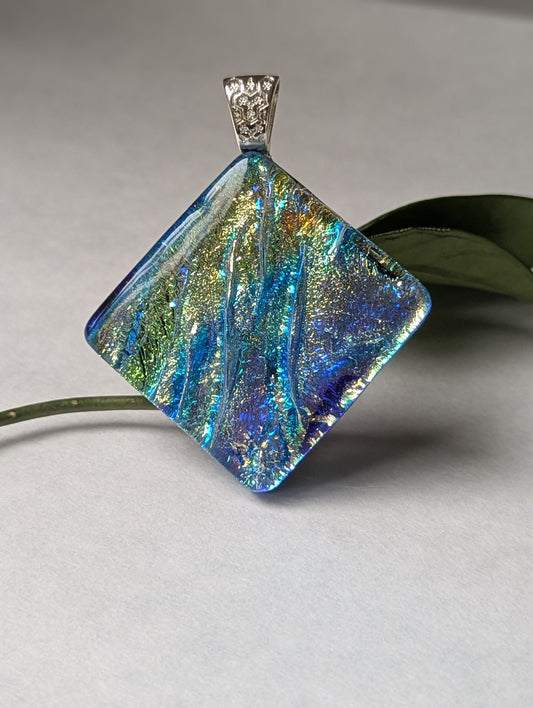 A blue iridescent fused glass pendant with a silver metallic clasp, displayed on a leaf.