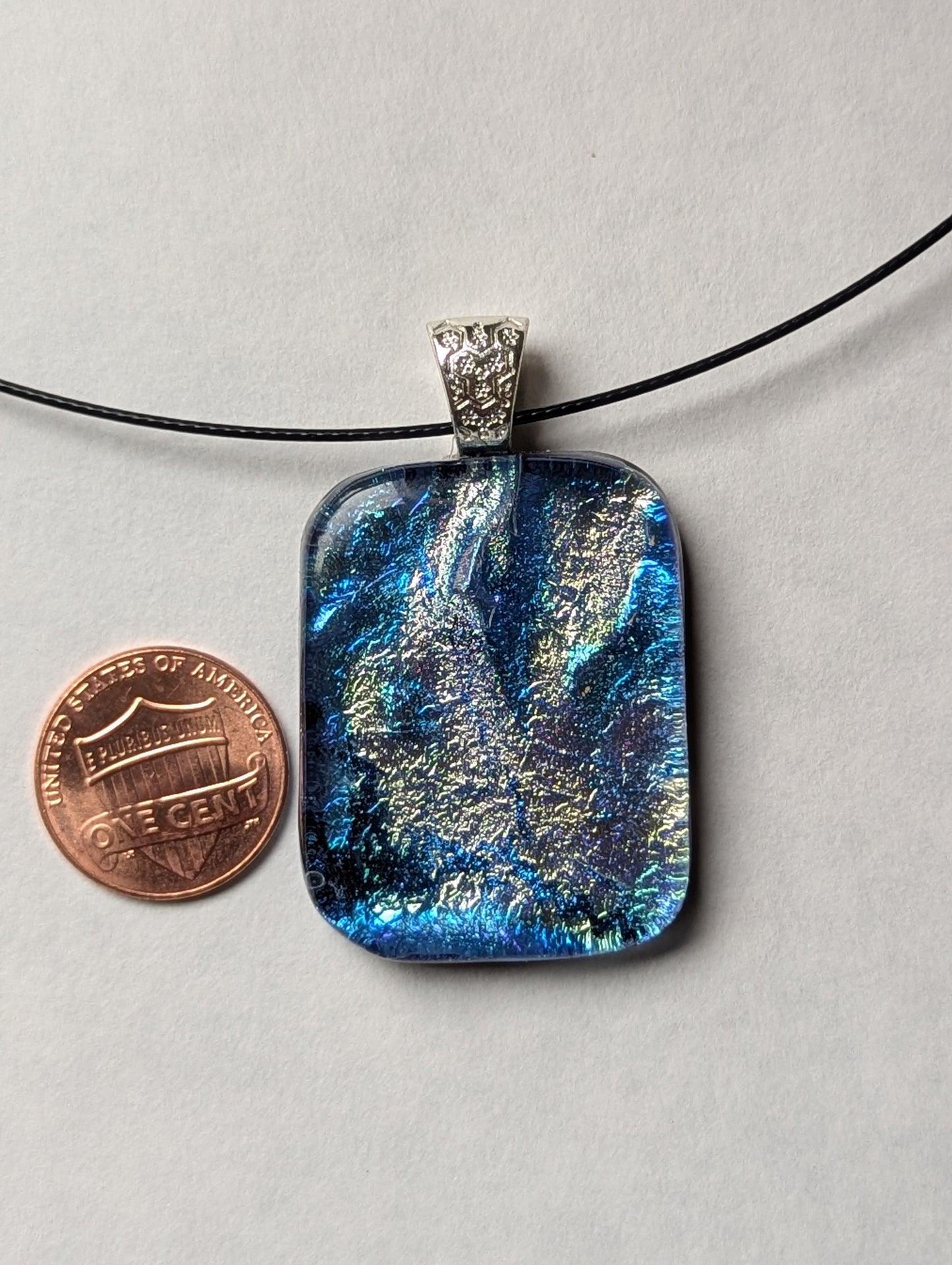 A blue and silver iridescent fused glass pendant with a silver metallic clasp, displayed on a black stainless steel necklace next to a penny for size reference.