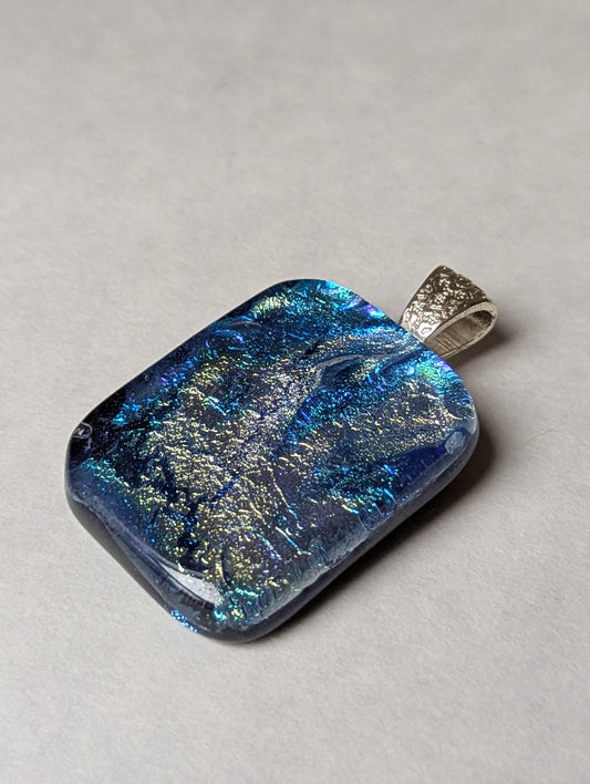 A blue and silver iridescent fused glass pendant with a metallic clasp, displayed against a white background.
