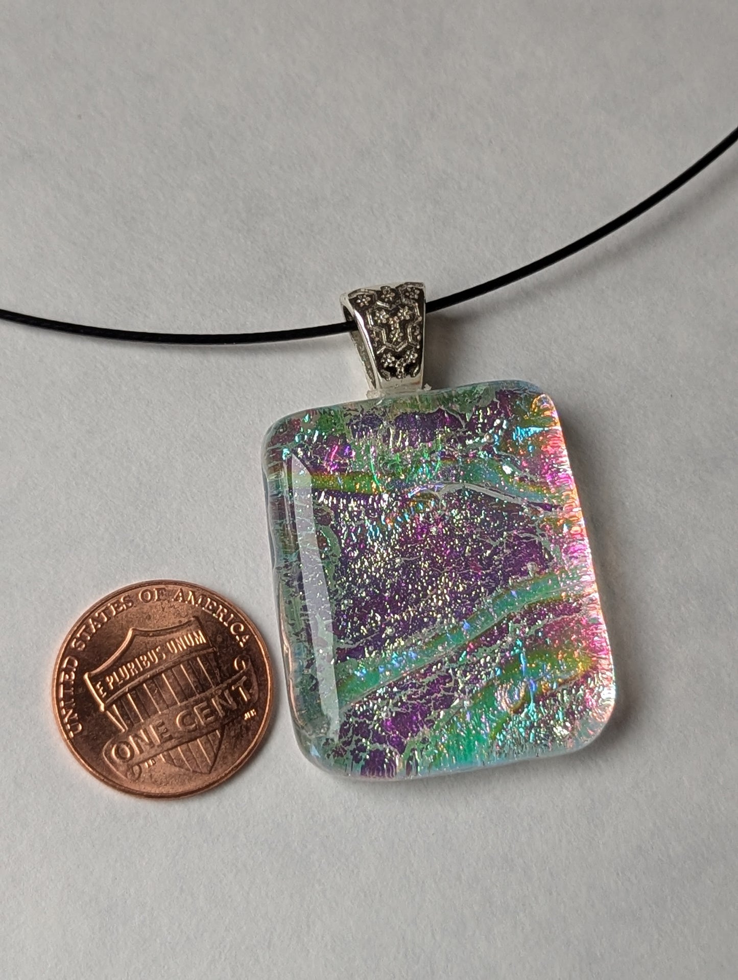 A colorful clear iridescent fused glass pendant with a silver metallic clasp, displayed on a black stainless steel necklace next to a penny for size reference.