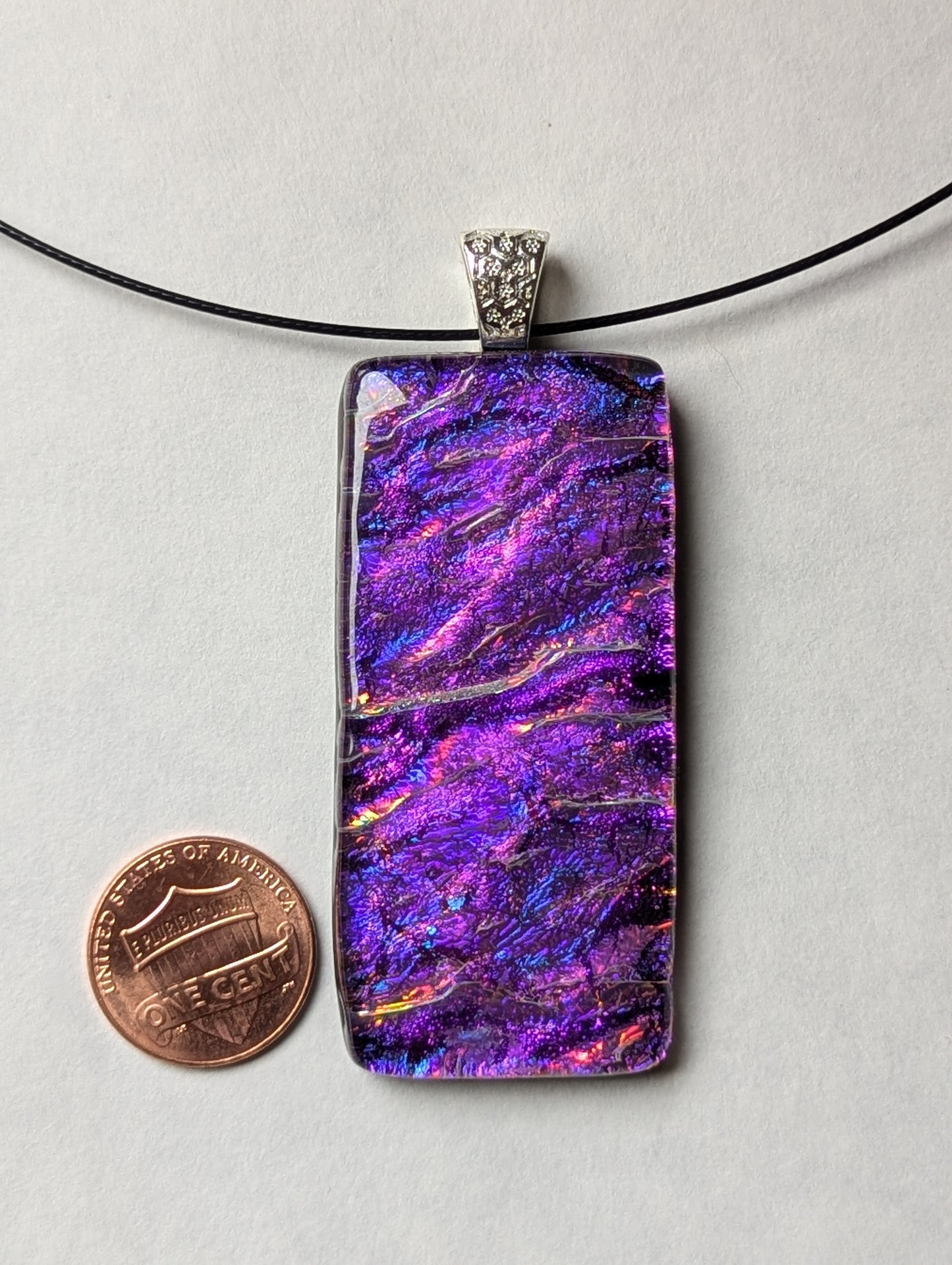 A purple iridescent fused glass pendant with a silver metallic clasp, displayed on a black stainless steel necklace next to a penny for size reference.