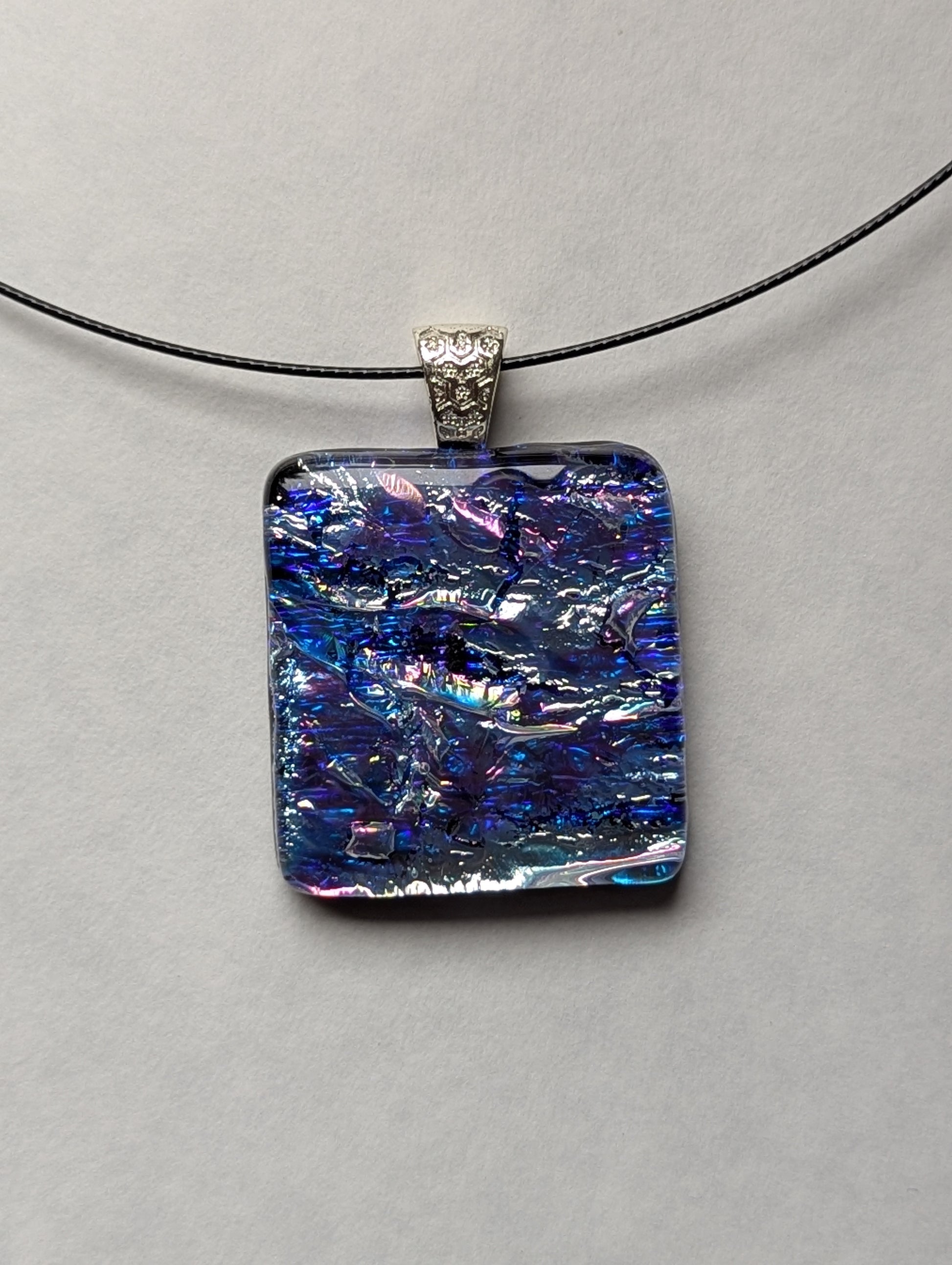 A blue and silver iridescent fused glass pendant with a metallic clasp, displayed on a black stainless steel necklace against a white background.