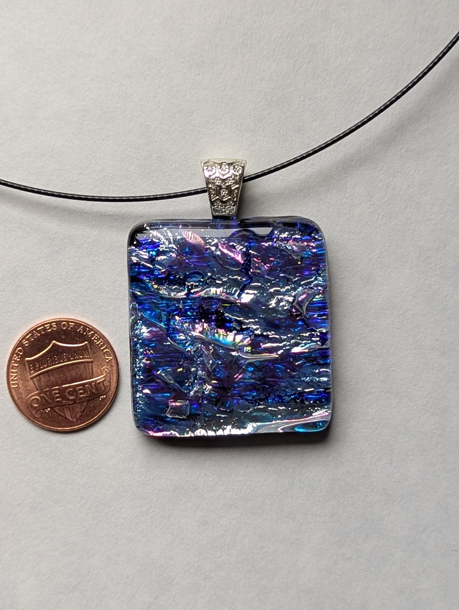 A blue and silver iridescent fused glass pendant with a silver metallic clasp, displayed on a black stainless steel necklace next to a penny for size reference.
