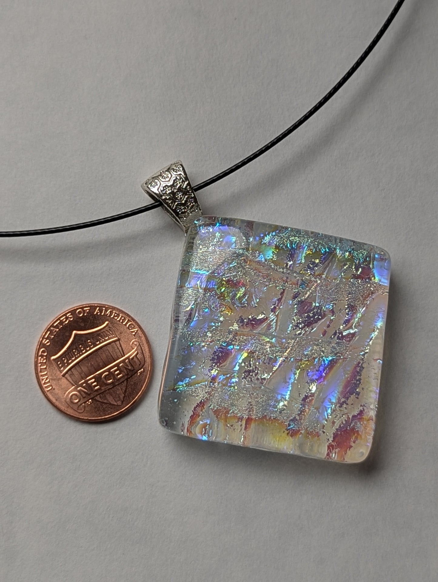 A colorful clear iridescent fused glass pendant with a silver metallic clasp, displayed on a black stainless steel necklace next to a penny for size reference.