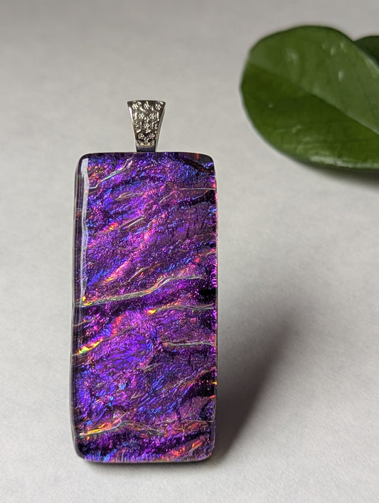 A purple iridescent fused glass pendant with a metallic clasp, displayed against a neutral background with a green leaf to the side.